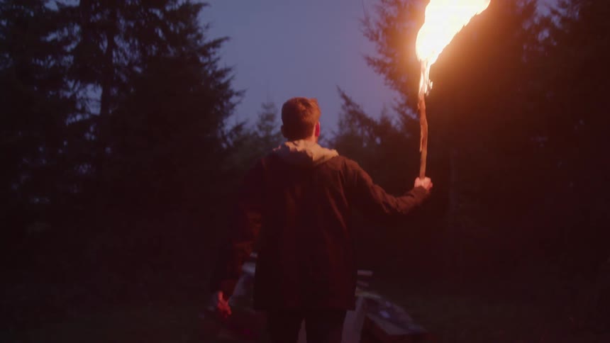 A person carrying a burning stick walks toward a campsite in a forest
