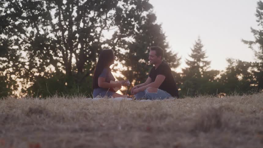 Couple sitting on the grass at sunset while she holds a ukulele
