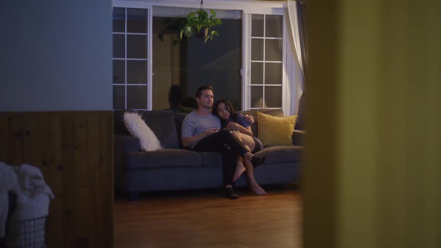 A happy couple is seen from behind sitting closely together on a couch relaxing at home in front of a window