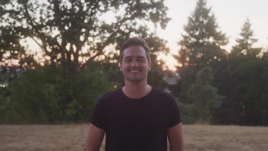 Man in a black tshirt smiles in an open field at sunset