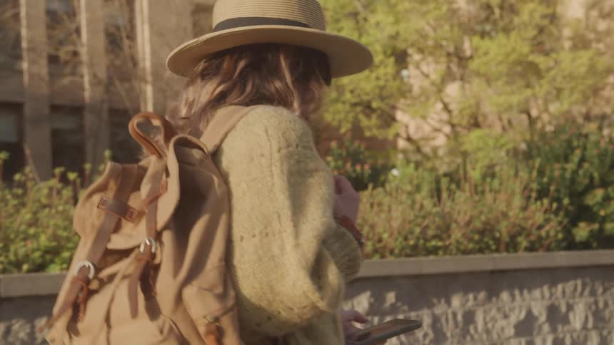 A person wearing a straw hat and a brown backpack is walking outdoors the person is also wearing a fluffy lightcolored sweater and holding a smartphone in one hand