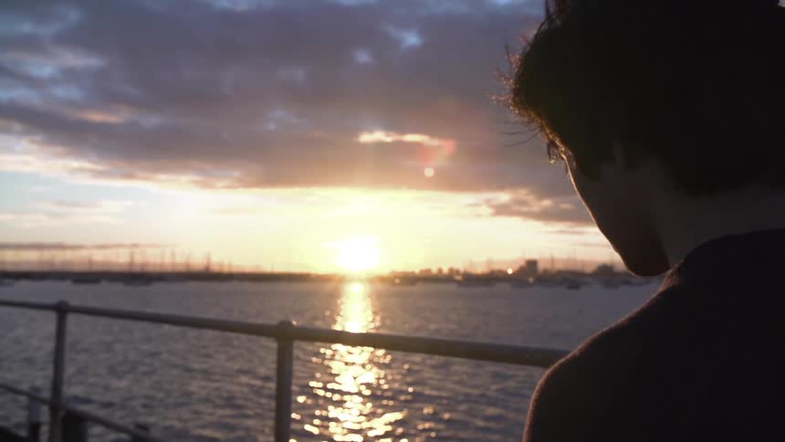 A man standing on a boardwalk watching the water at sunset