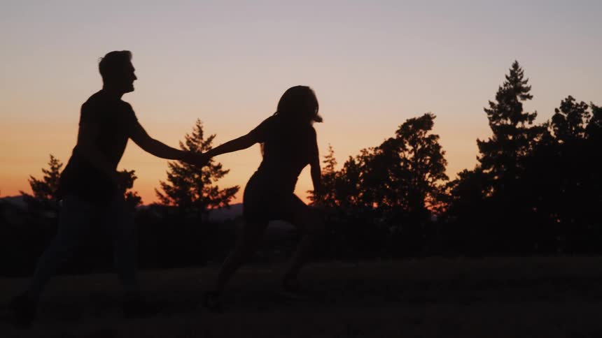 A man and a woman who are silhouetted by the sunset run hand in hand through a field with dark trees and mountains on the horizon