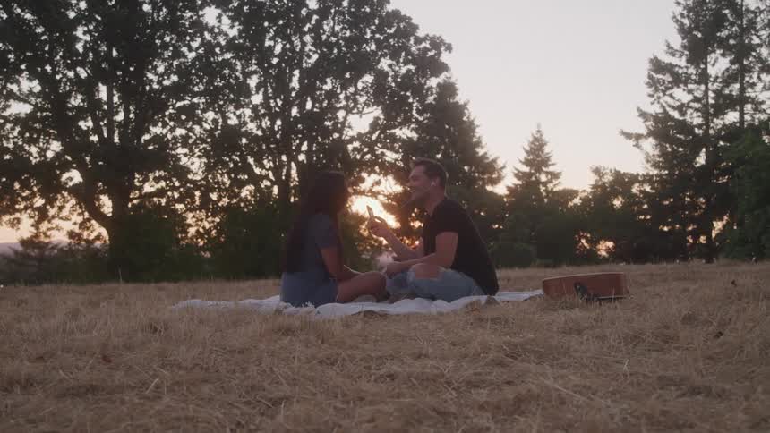 A couple sits on a grassland on a blanket having a fun time together as the sun sets