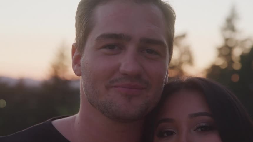A smiling man stands outdoors during golden hour with a womans face partially visible beside him