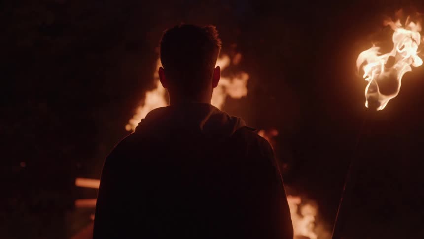 A man holds a flaming torch with a fire burning in front of him in the woods at night