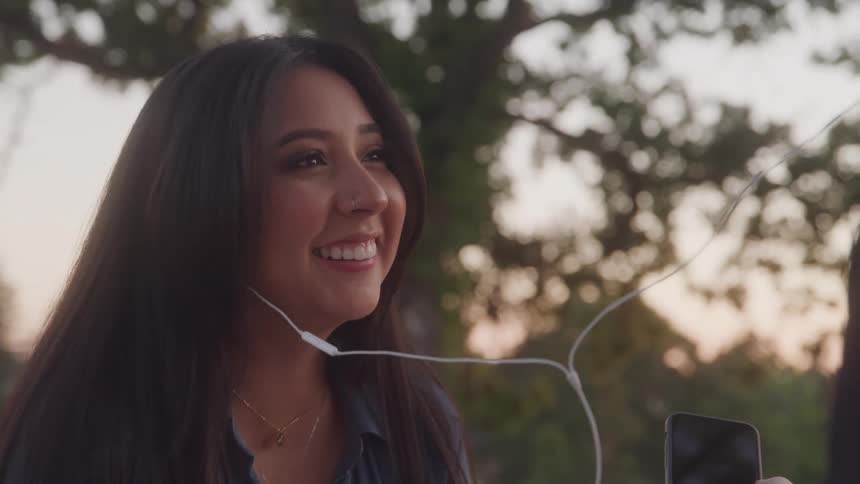 A happy couple shares a pair of headphones while looking at a phone with the woman smiling as the man talks or listens