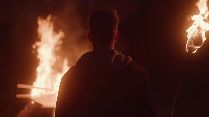 Man from behind watches a bonfire while holding a lit torch