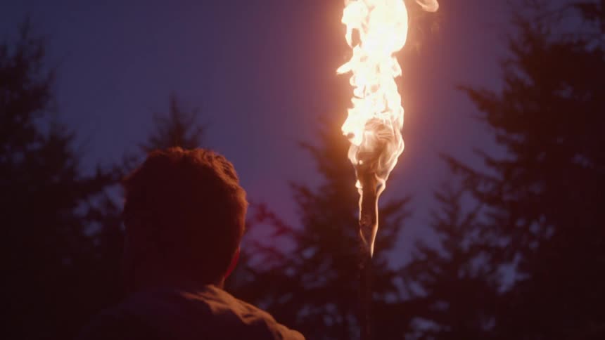 A man stands in the forest holding a flaming torch at night