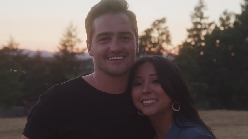 A smiling couple poses closely together outdoors during sunset