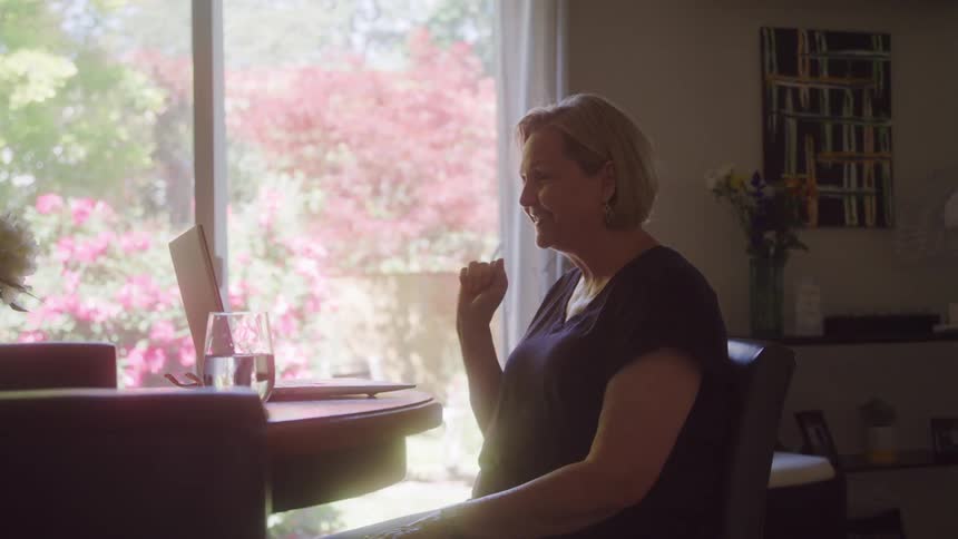 Smiling woman video chatting on a laptop seated at a table near a sunlit window