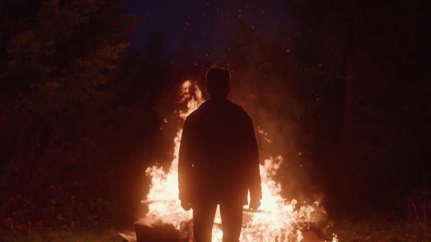 A man stands silhouetted against a blazing bonfire in the dark forest night