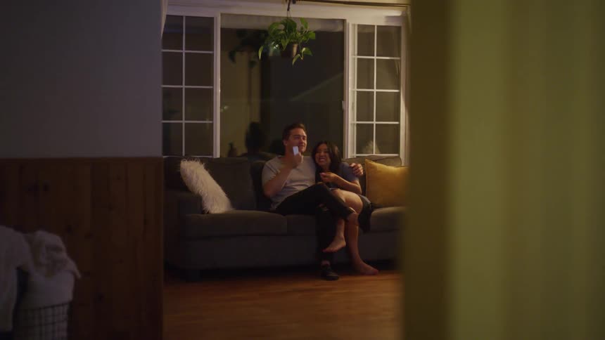 A man and woman sit close together on a couch at night smiling and watching something offscreen