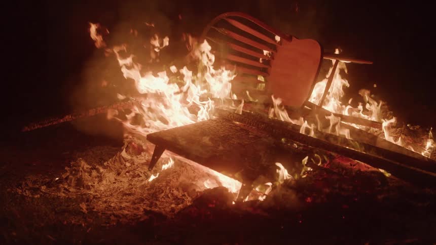 A chair and other pieces of wood are engulfed in flames burning in a bonfire at night the fire is bright and casts an orange glow on the surroundings