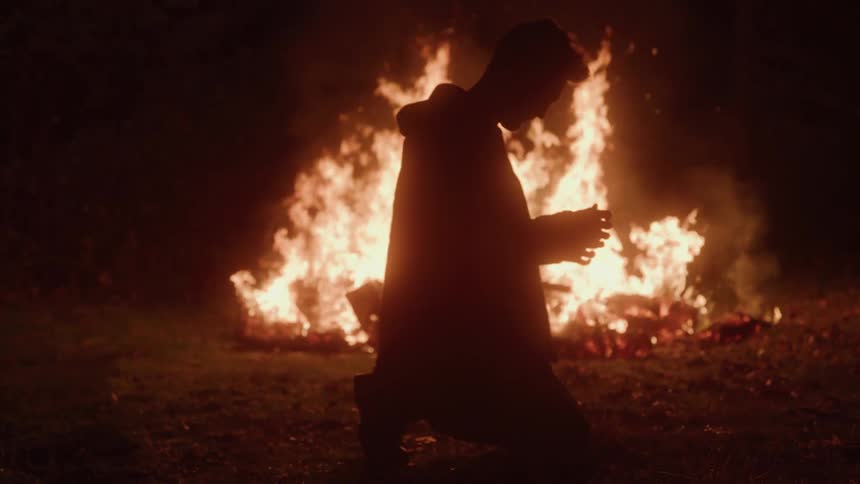 A young man wearing a long coat and pants kneels in front a large bonfire in the dark