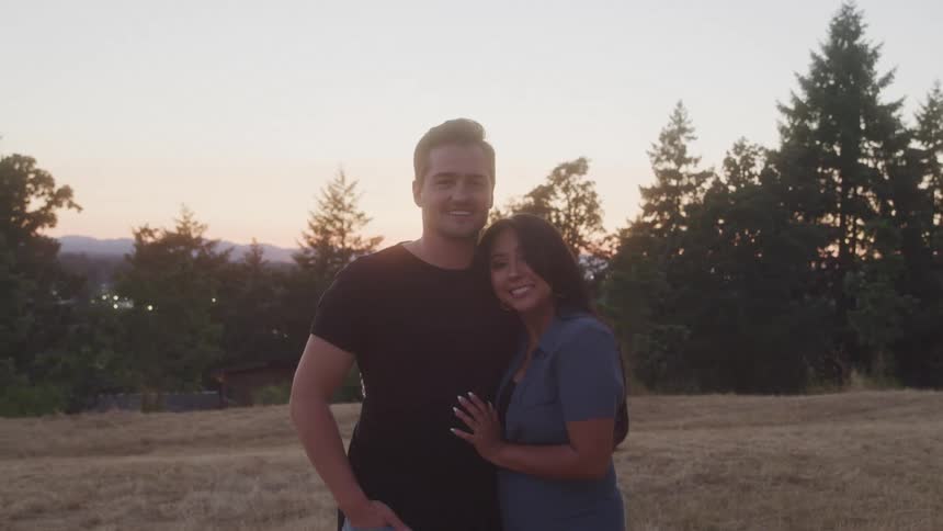 A man in a black shirt and a woman in a blue shirt stand together in a grassy field with trees and a sunset sky behind them both smiling directly at the camera