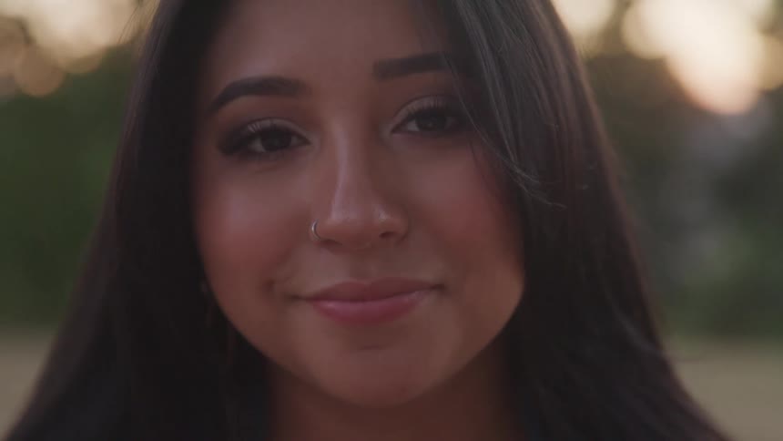 A young woman with long dark hair and a nose ring smiles softly in the golden day light