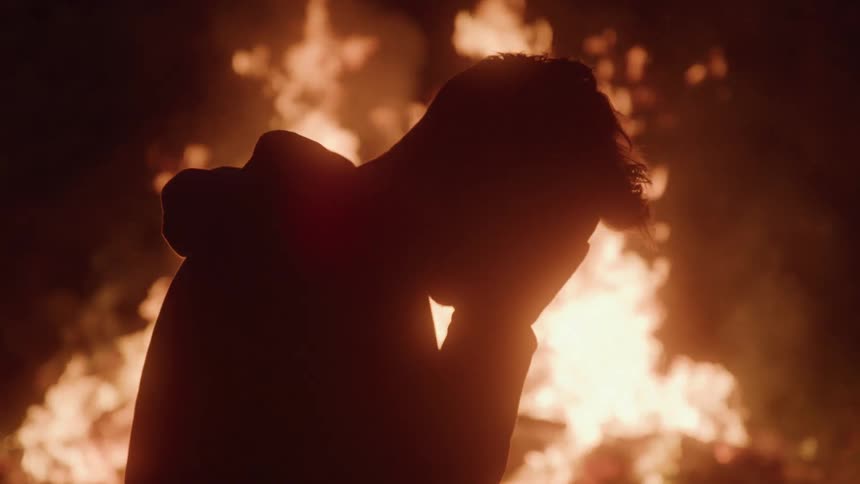 Silhouetted person covers their face in front of a large bonfire