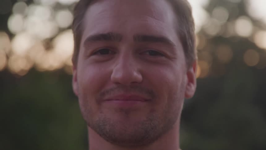 A closeup of a smiling man with light hair and a beard looking directly at the camera he is outdoors and the background is out of focus with bokeh from sunlight filtering through trees