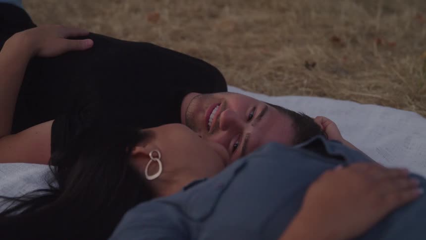 A couple lies on a blanket smiling at each other at midday on a grassland
