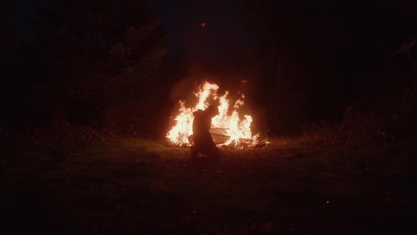 A man kneels with his back to a large bright bonfire in a dark wooded clearing