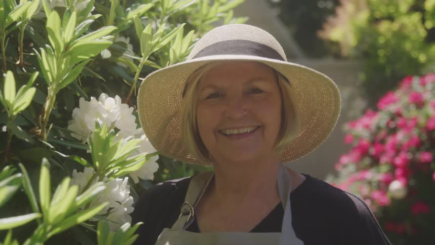 Older woman wearing a sunhat stands next to a flowering bush in the garden and smiles