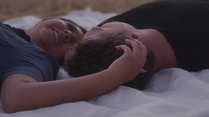 A woman and a man are lying down together on a lightcolored blanket the woman is smiling and looking up and her arm is draped over the man who is looking to the side