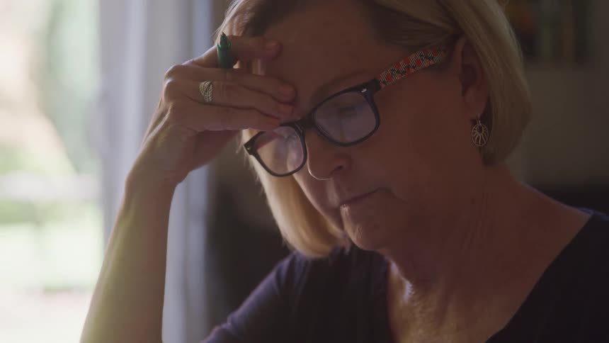 A woman with short blonde hair and glasses holds a pen to her forehead while reading