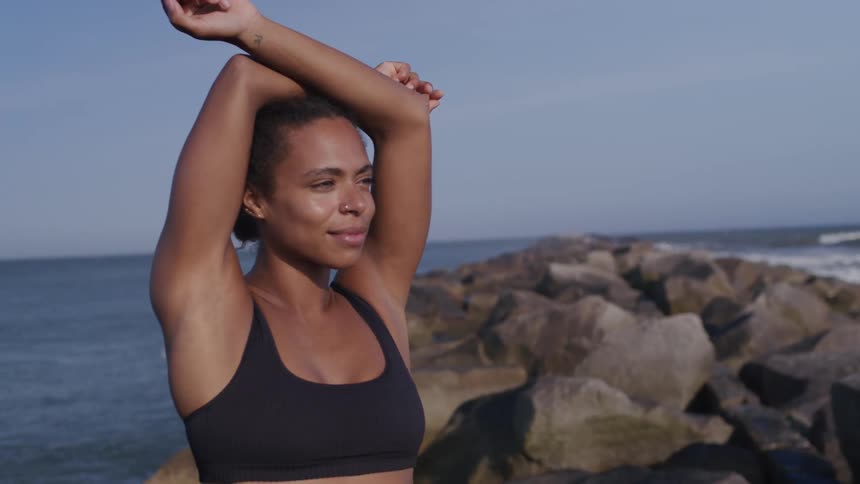 A woman wearing a black sports bra is doing a stretching pose with her arms raised over her head she is standing outdoors on a rocky jetty next to the ocean on a clear sunny day
