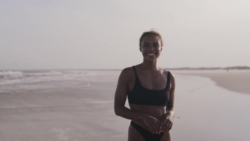 A woman in a black bikini walks on a sandy beach next to the ocean turns towards the camera and smiles