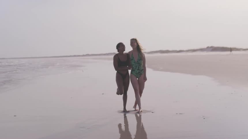 Two women in swimsuits pose together smiling at the shoreline