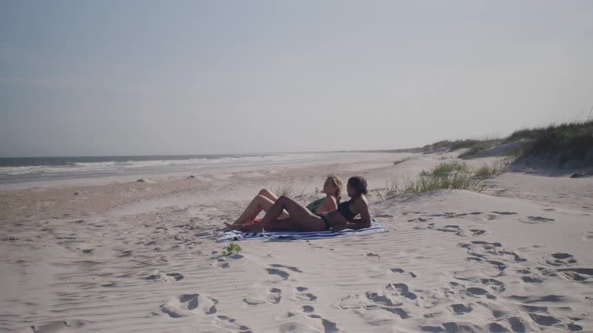 Two women relax on a towel laid out on the sand of a deserted beach