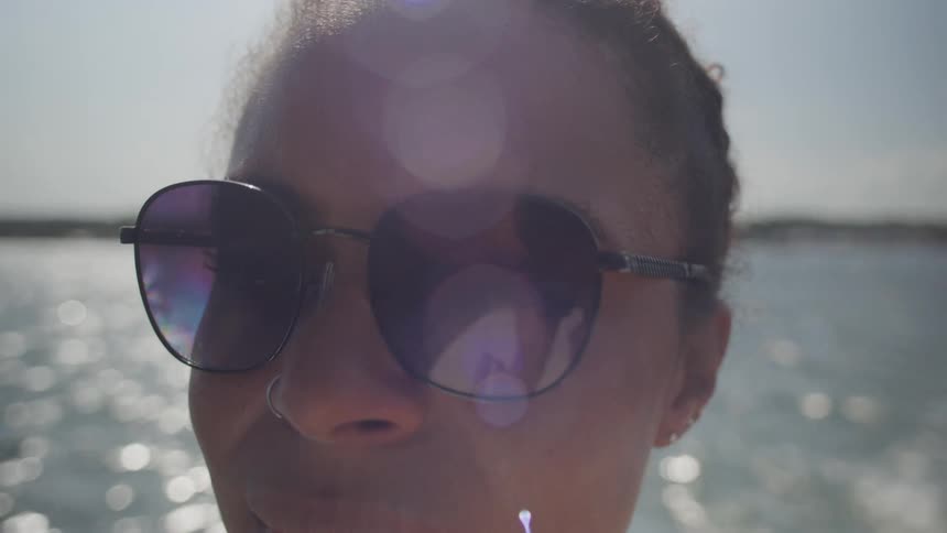 A woman wearing dark sunglasses stands near shimmering water with sunlight creating lens flares across her face