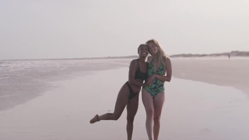 Two women in swimsuits walk arm in arm smiling at the beach shore