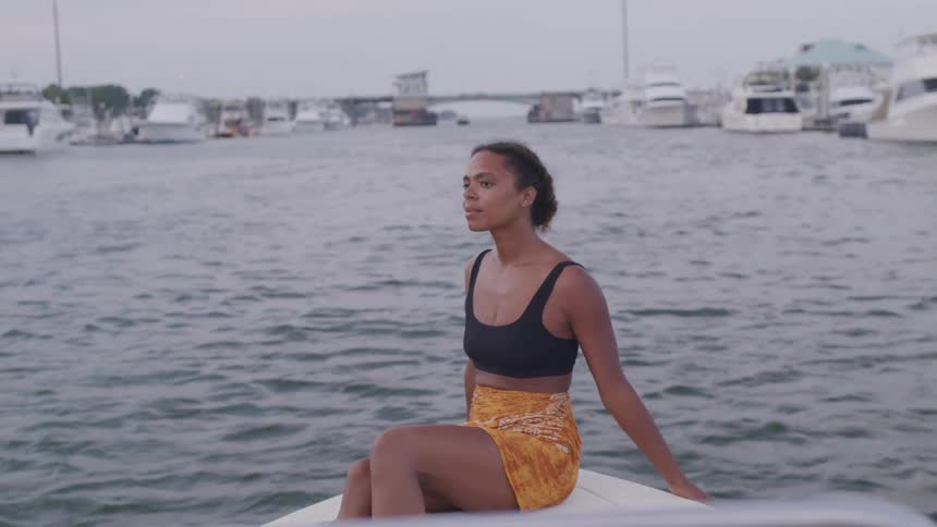 A woman sits calmly at the edge of a boat in a marina gazing out over the water