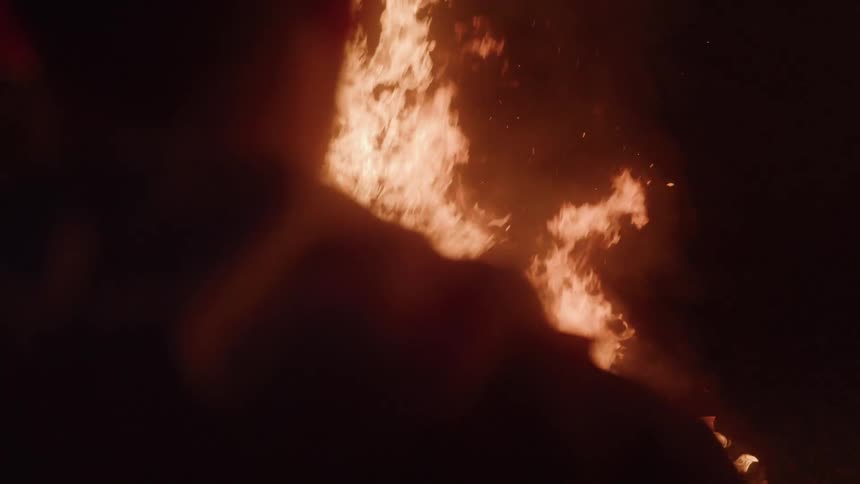A male stands near a glowing campfire seen from behind in the dark