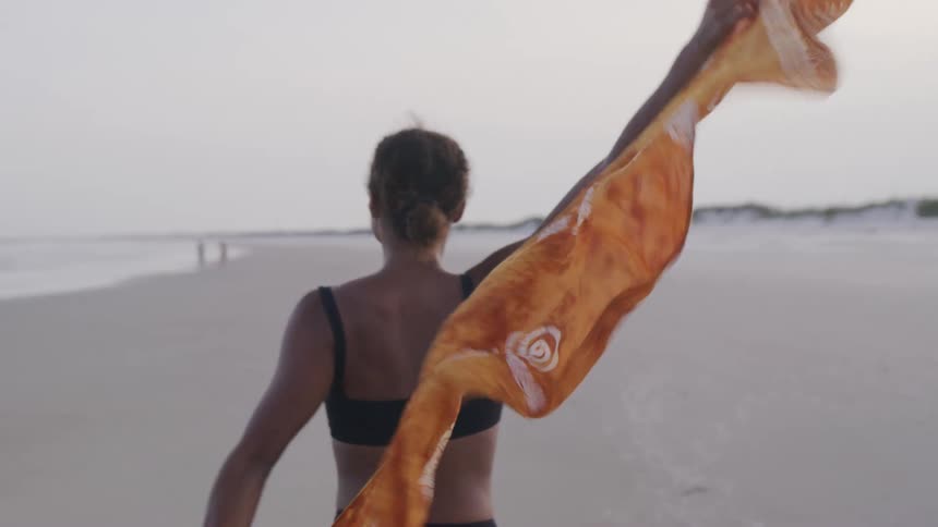 The video shows a woman walking on a beach at what appears to be dusk she is holding a large patterned orange scarf that billows behind her and she looks back over her shoulder with a joyful smile