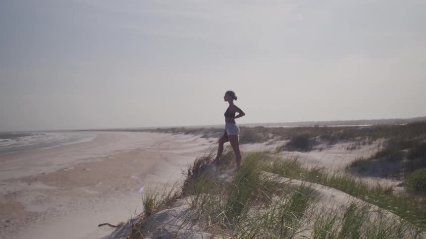 A woman stands on a sandy dune with tall grass looking out over a wide empty beach and the ocean on a bright day