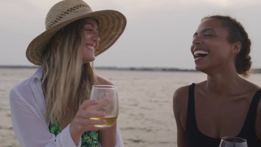 Two women enjoy drinks together on the beach at sunset