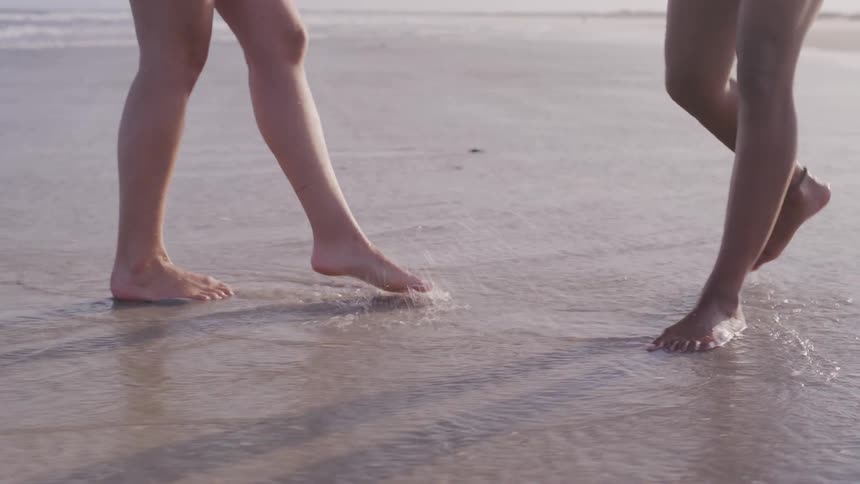 The video shows a closeup of the lower legs of two people walking on a wet sandy beach the waves gently wash over their feet as they stroll