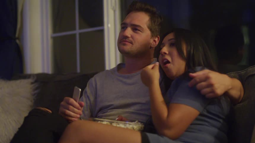 Couple snuggles on the couch at night sharing popcorn while watching tv