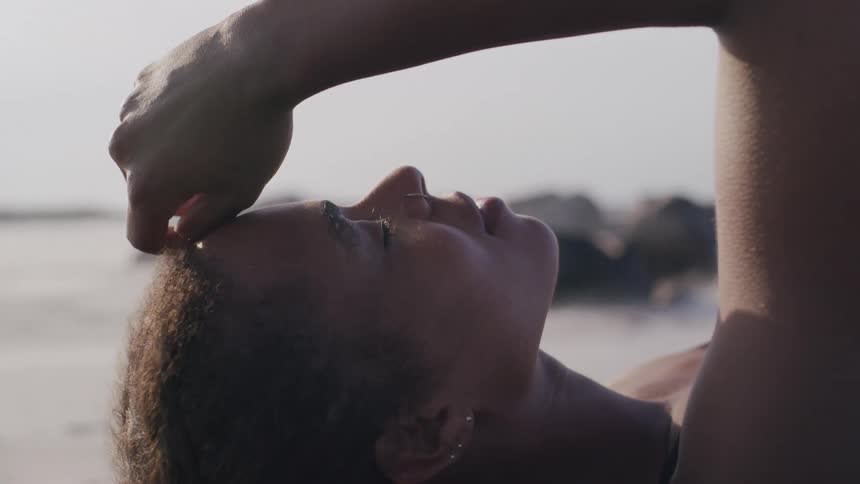 A woman lies on the beach with her arm resting on her forehead eyes closed in peaceful relaxation