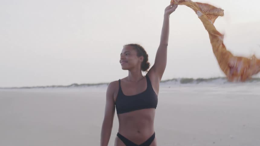 A woman wearing a black bikini and an orange patterned sarong stands on a sandy beach raising the sarong into the air and then letting it fall down