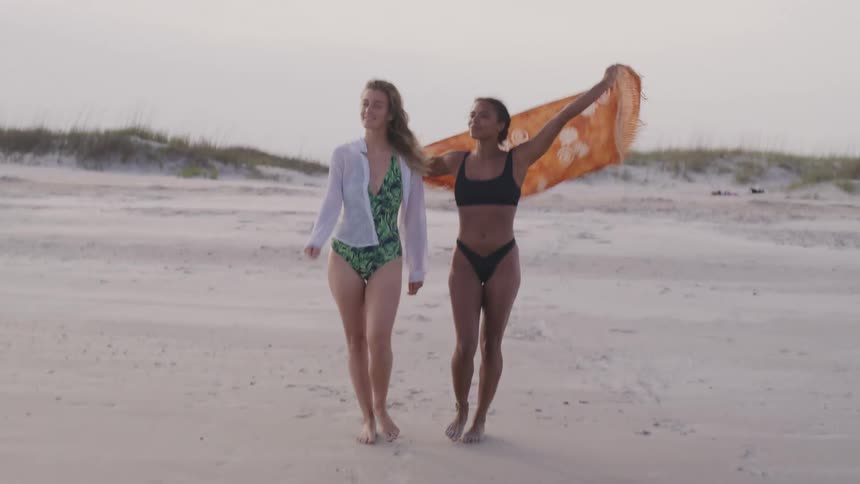 Two women walk along the beach sand with a scarf in the wind