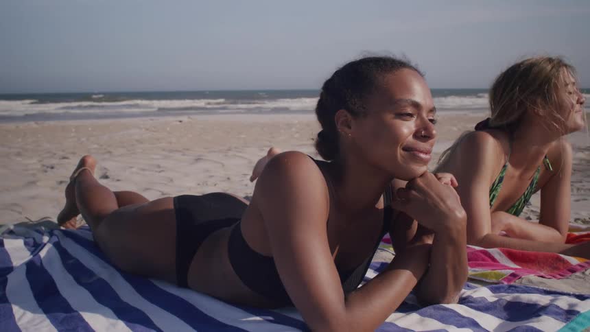 Two women lie on their stomachs on striped beach towels both smiling and relaxing near the coast