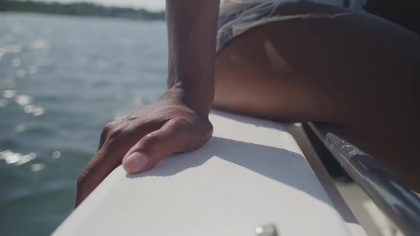 A persons hand grips the edge of a boat as sunlight sparkles on the water nearby
