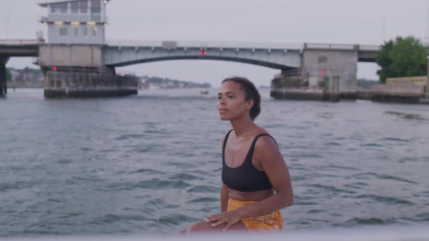 A woman sits calmly on a boat near a bridge at sunset