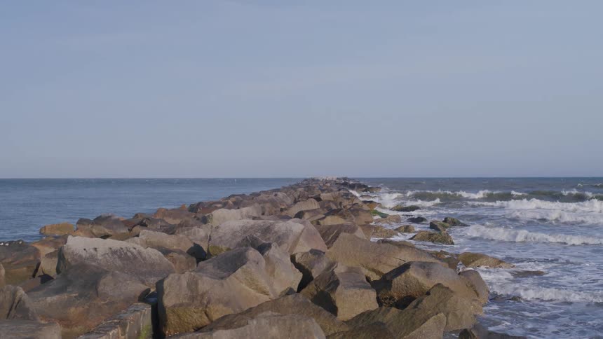 Waves from the ocean crash against a long rock jetty that stretches out to the horizon under a clear sky