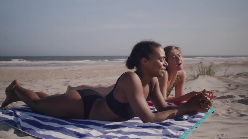 Two women in swimsuits lie on a striped towel at the beach smiling as they talk under the afternoon sun