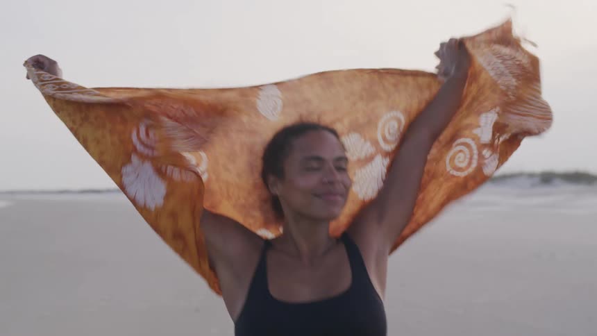 A woman in a black bikini walks on a sandy beach raising an orange patterned sarong above her head and then letting it fall down